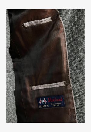 Pilka tekstūruota medžiaga viduje su dviem kišenėmis, turinti tamsų pamušalą ir audinio etiketę su užrašu "Shetland, Woven in England, Since 1837."