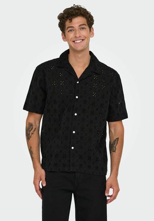 Chemise noire à manches courtes avec un tissu texturé et des découpes en forme de diamant, ornée de boutons blancs, associée à un pantalon noir.