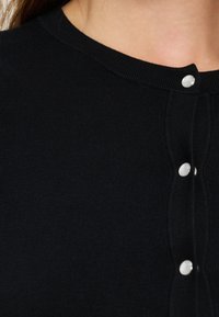 Μαύρο πλεκτό ένδυμα με στρογγυλή λαιμόκοψη και κάθετη σειρά από λαμπερές λευκές κουμπιά στο μπροστινό μέρος, εμφανιζόμενο στο πάνω μέρος του στήθους και του λαιμού.