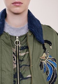 Personne portant une veste verte avec un col texturé bleu et une couche intérieure camouflage, ornée d'une broderie détaillée de cygne bleu et noir sur la poitrine.