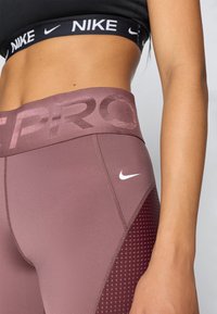 Leggings Nike rose et bordeaux avec une taille haute, présentant des accents en mesh et un logo blanc. Texture lisse avec un design ajusté.