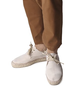 Zapatos de lona beige claro con punta redonda, diseño de cordones y suela de cuerda de yute. Combinados con pantalones marrones, mostrando materiales simples y naturales.