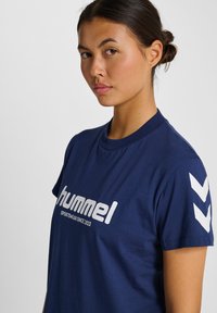 Mörkblått bomullströja med vit logotyp "hummel" och text. Kortärmad med kontrasterande vita chevron-accenter på axlarna.