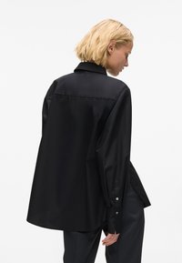Fekete, oversize ing sima anyagból, klasszikus gallérral, gombos mandzsettával és finoman fénylő, aszimmetrikus hemrésszel.
