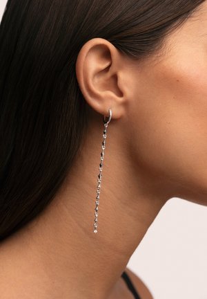 Pendiente de aro de plata con una cadena larga que combina eslabones brillantes y texturizados, y un detalle de piedra preciosa transparente al final.