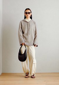 Sudadera gris con cordón, de corte oversized. Pantalones cargo marfil, bolso negro holgado y gafas de sol negras. Texturas suaves, diseño minimalista.