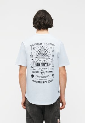 Personne aux cheveux noirs portant un t-shirt blanc avec des textes graphiques noirs et des symboles, dont un œil dans un triangle et des crânes au dos.
