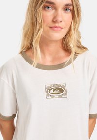 Witte T-shirt met korte mouwen, olijfgroene afwerking, met een geometrisch grafisch ontwerp in bruin op de borst. Zachte textuur.