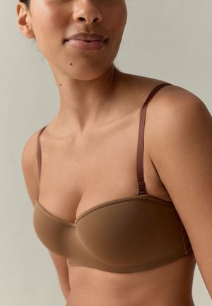 Vrouw draagt een bruine strapless beha met dunne verstelbare bandjes, schouder en bovenborst zichtbaar tegen een neutrale achtergrond.