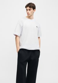 Νεαρός άνδρας που φοράει λευκό φαρδύ T-shirt και μαύρο παντελόνι με φαρδιά μπατζάκια, στέκεται με τα χέρια στις τσέπες, κοιτώντας προς τα αριστερά του.