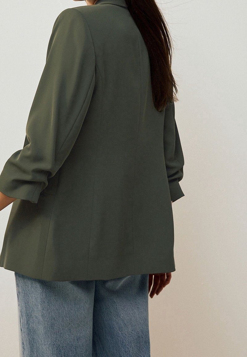 Personne avec de longs cheveux foncés portant un blazer vert olive aux manches retroussées et un jean large bleu clair, debout de profil.