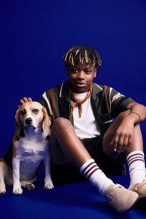 A boy sits beside a beagle on a blue background. He wears a multi-colored striped shirt, black shorts, and cream shoes with red accents.