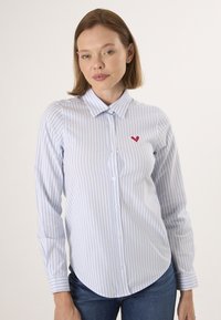 Camicia button-up a righe azzurro chiaro e bianco con orlo curvo e ricamo di un cuore rosso sul taschino sinistro. Maniche lunghe e colletto classico.