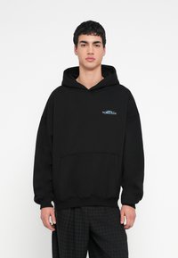 Hanorac negru cu buzunar frontal, având un logo brodat albastru. Fabricat dintr-un material moale, cu croială lejeră și glugă reglabilă cu șnur.