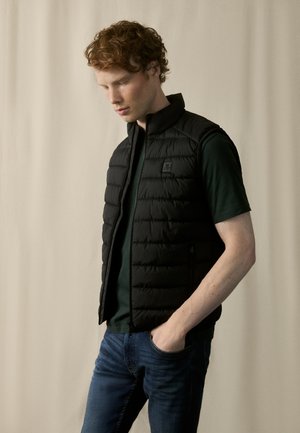 Chaleco acolchado negro con cuello alto y cierre de cremallera, texturizado con costuras horizontales, llevado sobre una camiseta verde y unos jeans azules.