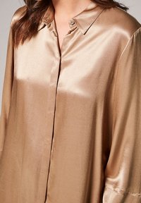 Camicia button-up in satin marrone chiaro con colletto, caratterizzata da una texture liscia e una vestibilità ampia. Include un dettaglio con i bottoni visibile sul colletto.