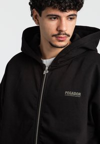 Sort zip-up hoodie lavet af bomuld, med en stor hætte, sølv lynlås og et logo på brystet. Enkel tekstur, minimalistisk design.