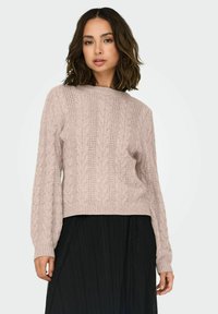 Pull en maille torsadée rose clair avec un col rond, des manches longues et un ourlet côtelé, associé à une jupe plissée noire.