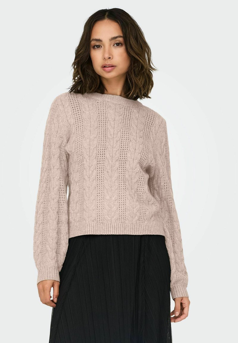 Pull en maille torsadée rose clair avec un col rond, des manches longues et un ourlet côtelé, associé à une jupe plissée noire.