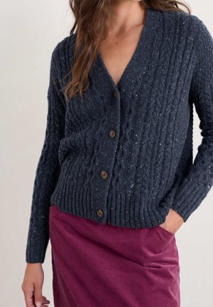 Femme portant un cardigan boutonné bleu foncé en maille torsadée et une jupe taille haute magenta foncé, une main dans la poche.