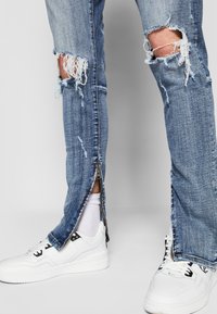 Jean bleu déchiré avec des bords effilochés, comportant une fermeture éclair sur les jambes inférieures. Associé à des baskets blanches et des chaussettes blanches visibles.