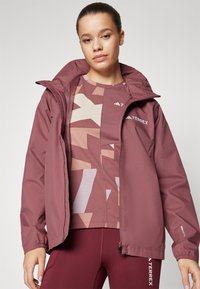 Chaqueta impermeable color marrón oscuro con capucha ajustable, que presenta puños elásticos y bolsillos laterales. Parte superior deportiva con estampado en tonos de rosa y marrón.