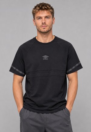 Uomo con capelli corti e ricci castani che indossa una maglietta nera Umbro con logo sul petto e bordi delle maniche a fantasia, mani nelle tasche dei pantaloni grigi.