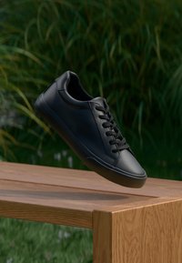 Schwarze Ledersneaker mit glatter Textur, runder Zehenpartie und sechs Ösen. Gummisohle und minimalistisches Design, präsentiert auf einer Holzoberfläche.