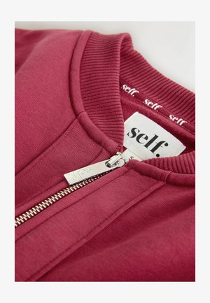 Vínově červená bunda na zip s žebrovaným límcem, stříbrným zipem a textilní štítkem s nápisem „self.“ v černé barvě. Měkká textura, neformální design.