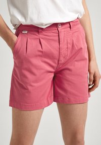 Shorts in cotone rosa con pieghe frontali e chiusura a bottone, dotati di tasche laterali e orlo cucito. Logo applicato sulla vita.
