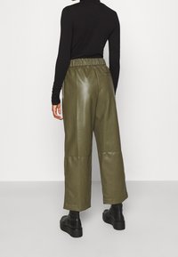 Pantaloni a gamba larga in pelle verde oliva con vita alta, caratterizzati da una superficie liscia e due tasche posteriori. Indossati con un top nero a maniche lunghe.