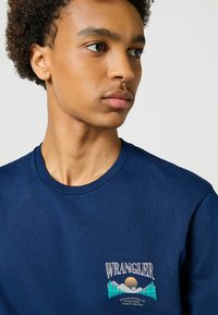 Mørkeblå bomulds t-shirt med et broderet logo med bjerge og solnedgangsdesign på venstre bryst. Klassisk crew neck.