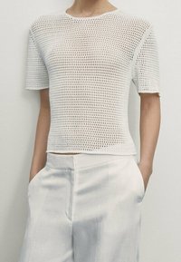 Vit stickad topp med korta ärmar, som har en öppen vävstruktur och en klassisk rundhalsringning, kombinerad med ljusa byxor.