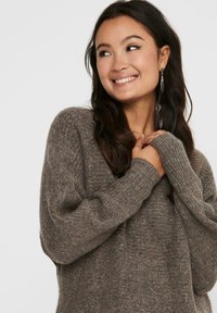 Pull brun texturé avec un col en V profond, coupe ample et manches longues. Le tissu semble doux et confortable, avec des détails côtelés aux manches.