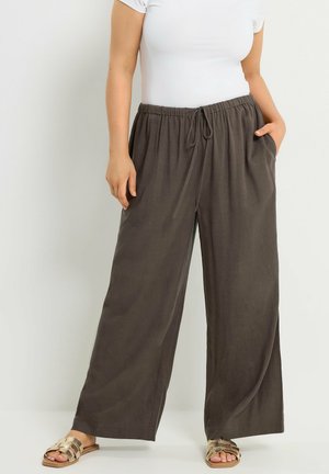 Person trägt lockere, dunkelgraue Kordelzug-Hose, weißes kurzärmeliges Oberteil und offene Sandalen und steht mit einer Hand in der Tasche.