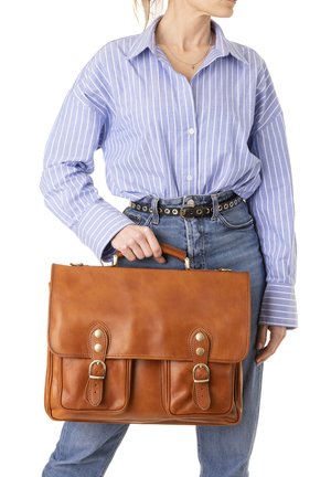 Donna che indossa una camicia a righe blu e jeans tiene una borsa ventiquattrore in pelle marrone per la maniglia su sfondo bianco.