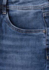 Poche de jean en denim bleu avec coutures visibles, passants de ceinture et petit rivet en métal dans le coin supérieur droit de la poche.