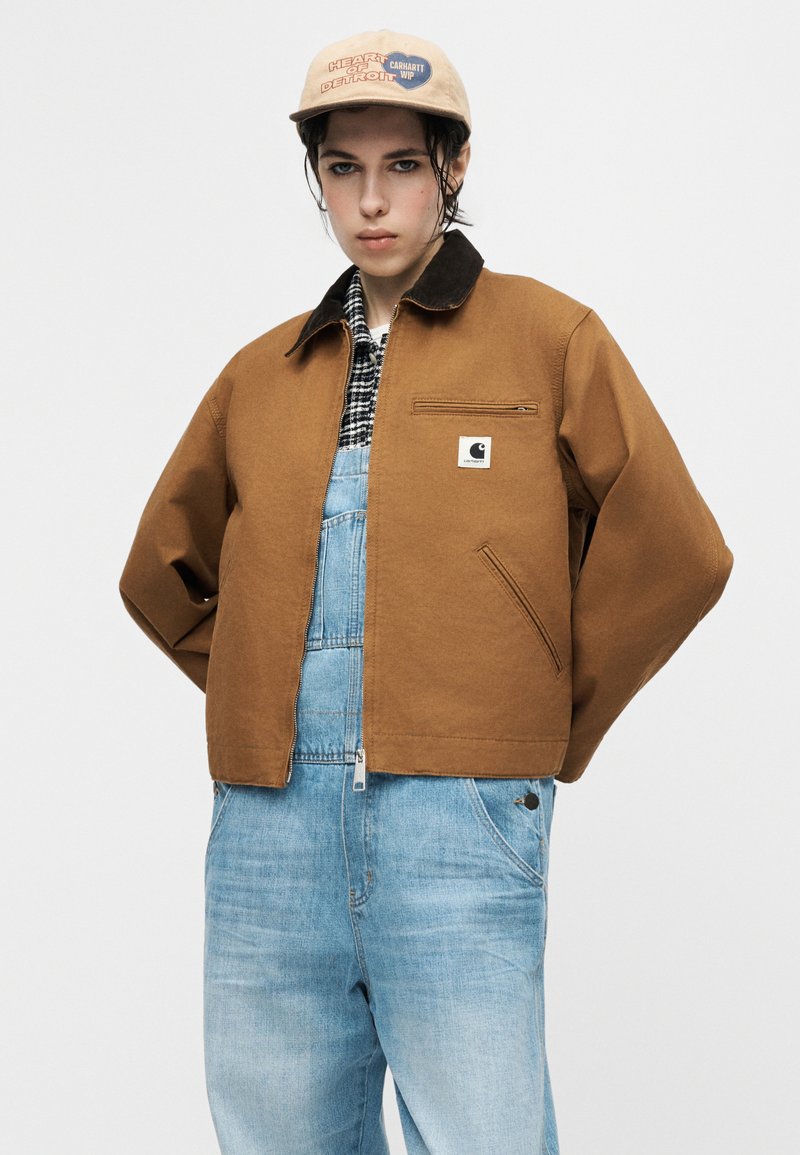 Person wearing beige cap, brown Carhartt jacket over black-and-white checkered shirt and light blue denim overalls, standing with hands behind back.