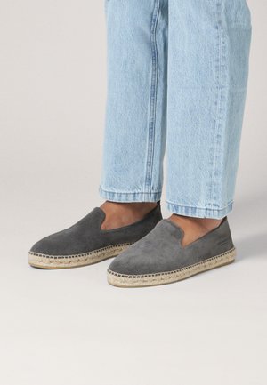 Person, die hellblaue Jeans und dunkelgraue Espadrilles aus Wildleder mit Jutesohlen trägt, steht vor einem schlichten hellen Hintergrund.