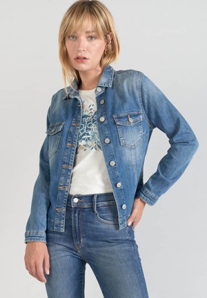 Veste en denim délavé clair avec un devant à boutons, deux poches poitrine et un col classique. Portée par-dessus un t-shirt à motifs et associée à un jean.