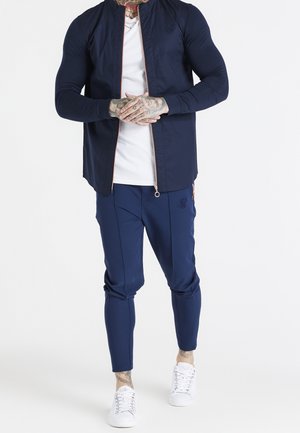 Homme portant une veste zippée bleu marine, un pantalon de jogging assorti, un t-shirt blanc et des baskets blanches, mains jointes devant, tatouages visibles sur les mains et le cou.