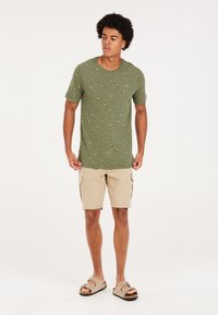 Alyvuogių žalia marškinėliai su mažomis kelių spalvų raštais, suderinti su smėlio spalvos krovinių šortais. Modelis dėvi rusvas šlepetes.