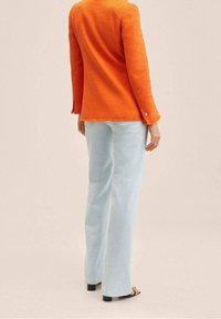 Oranžová texturovaná bunda s dlouhými rukávy, s třepeným lemem a detaily na knoflících, spárovaná se světle modrými kalhotami širokého střihu. Viditelné černé sandály.