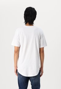 T-shirt blanc à manches courtes avec un col rond, présentant une coupe droite et un ourlet légèrement arrondi. Porté avec un jean bleu.