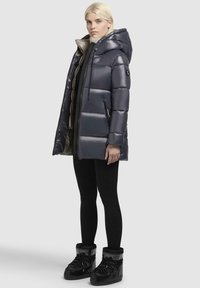 Gevoerde marineblauwe jas met capuchon en ritssluiting, met een glanzende afwerking, zwarte accenten en zijzakken, gecombineerd met zwarte sneeuwlaarzen.