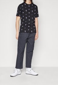 Henkilö, joka käyttää mustaa T-paitaa, jossa on violetteja Nike-logoja ja tekstiä, tummanharmaita housuja, valkoisia sukkia ja valkoisia Nike-tennareita mustalla swooshilla.