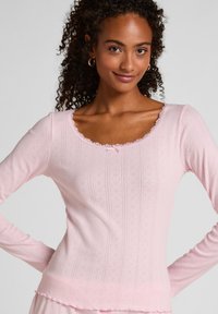Rosa langärmliges Oberteil mit einer wellenförm ausgeschnittenen Halslinie, Spitzenbesatz, strukturiertem Muster und einer kleinen Schleife in der Mitte. Weicher, leichter Stoff.
