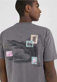 Camiseta de algodón gris con un gran gráfico de cocodrilo negro y estampas vintage coloridas en la parte posterior, con una textura desgastada.