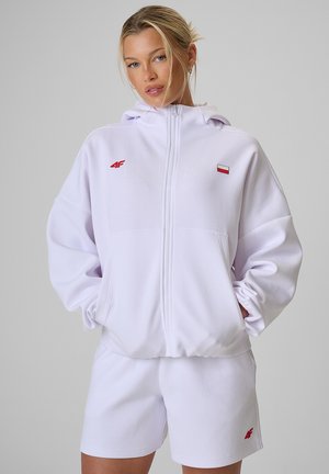 Blondynka ubrana w białą bluzę z kapturem zapinaną na suwak z małą flagą i logo, w połączeniu z białymi szortami na jednolitym szarym tle.