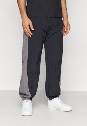 Homme portant un pantalon de sport noir et gris avec des poignets élastiques et des baskets blanches Champion, se tenant devant un fond uni.